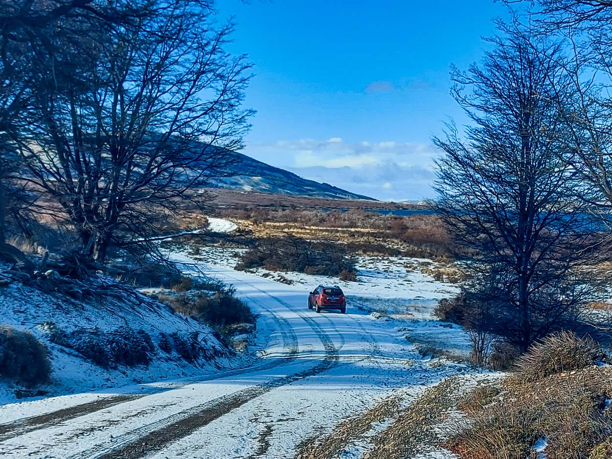 Un camino nevado entre árboles pelados, con una camioneta roja pequeña a lo lejos y las huellas marcadas en la nieve